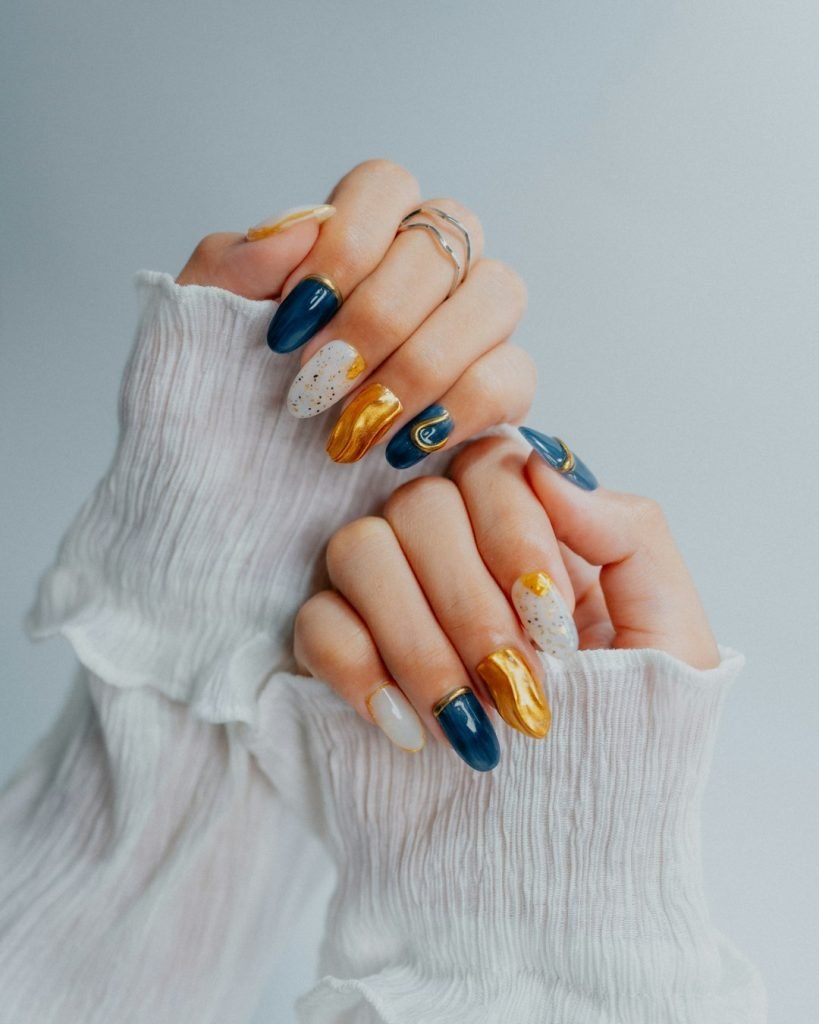 Close-up shot of hands with blue and gold nail art, featuring glossy finishes, speckled patterns, and a creative color mix.